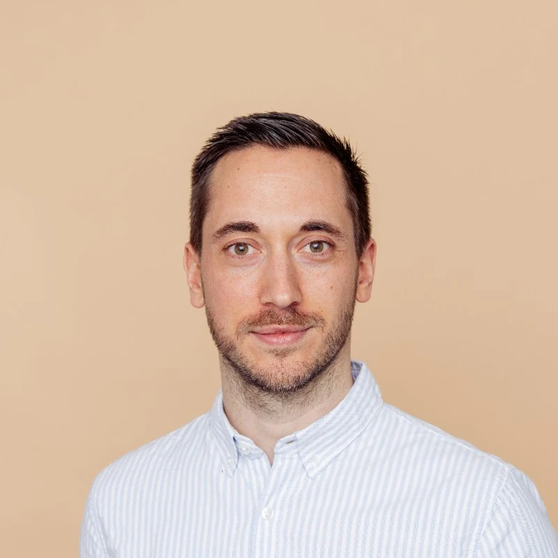 A man with short dark hair, light facial hair, wearing a light blue and white striped button-up shirt, smiling softly against a beige background.